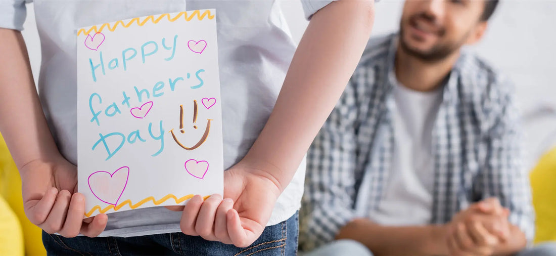 Image of a dad and child, the child is standing in front of dad holding a handmade card saying Happy Father's Day