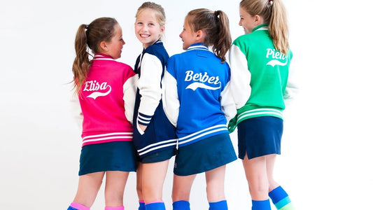 4 girls wearing varsity jackets printed with their names on the back. One wears pink, the others wear navy, blue and green.