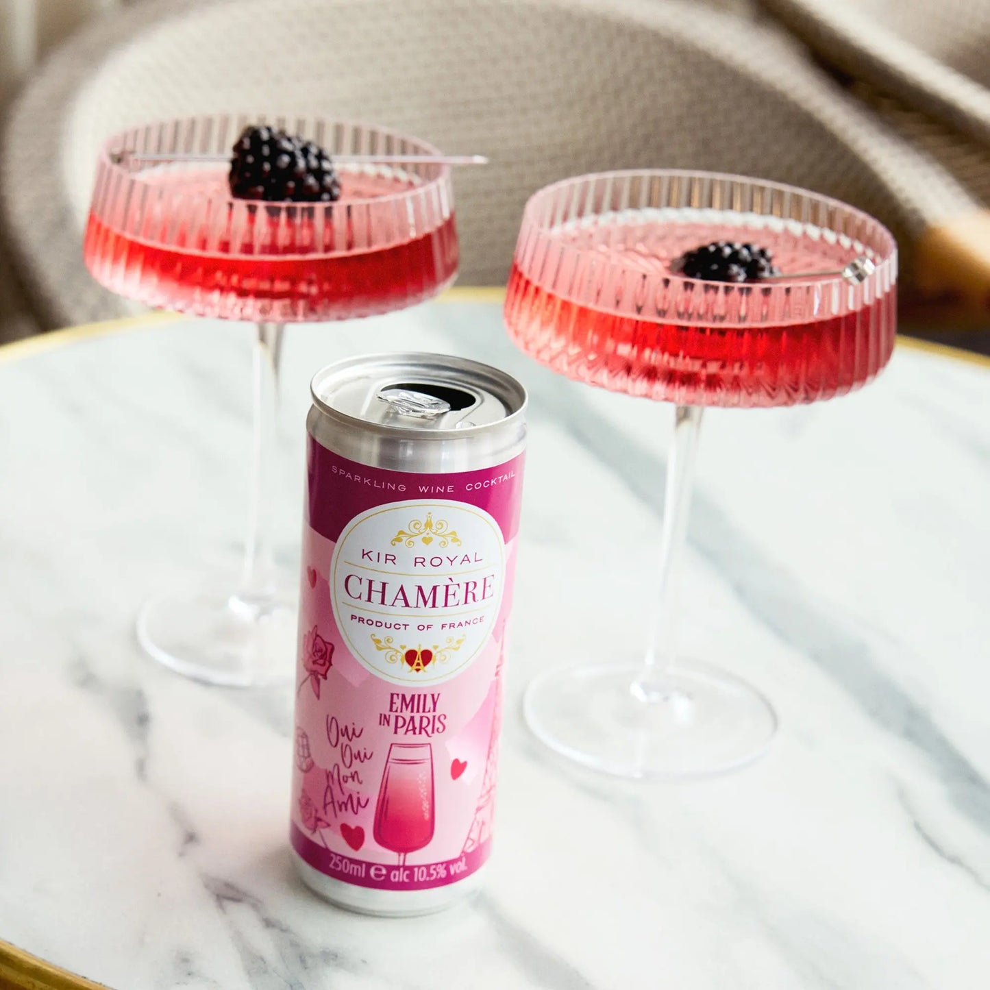 Can of Kir Royal Chamère wine cocktail in different shades of pink. With two champagne saucers filled with the cocktail with blackberry inside.