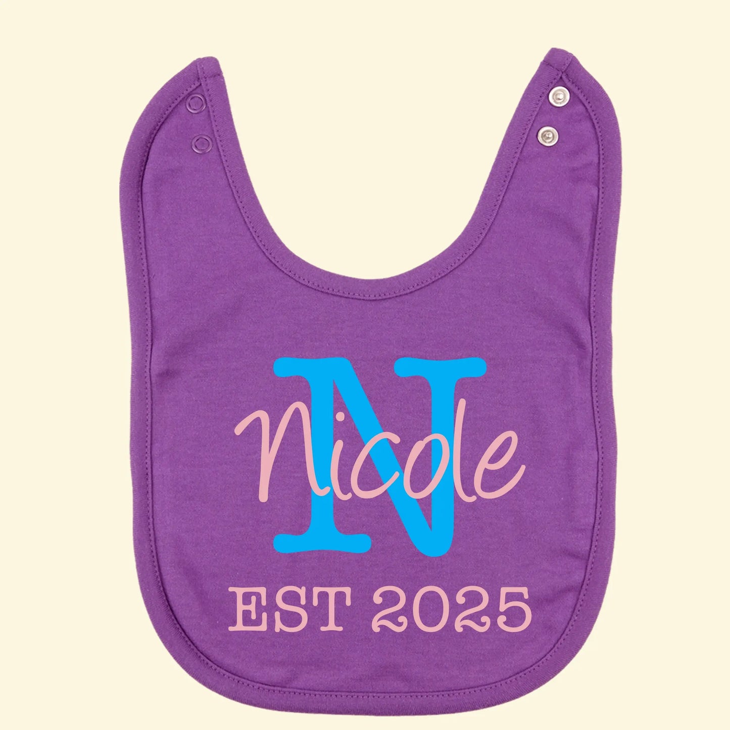 Purple bib printed with when the wearer was established, Nicole EST 2025 with the initial N behind. Printed in light pink for the text and blue for the initial.