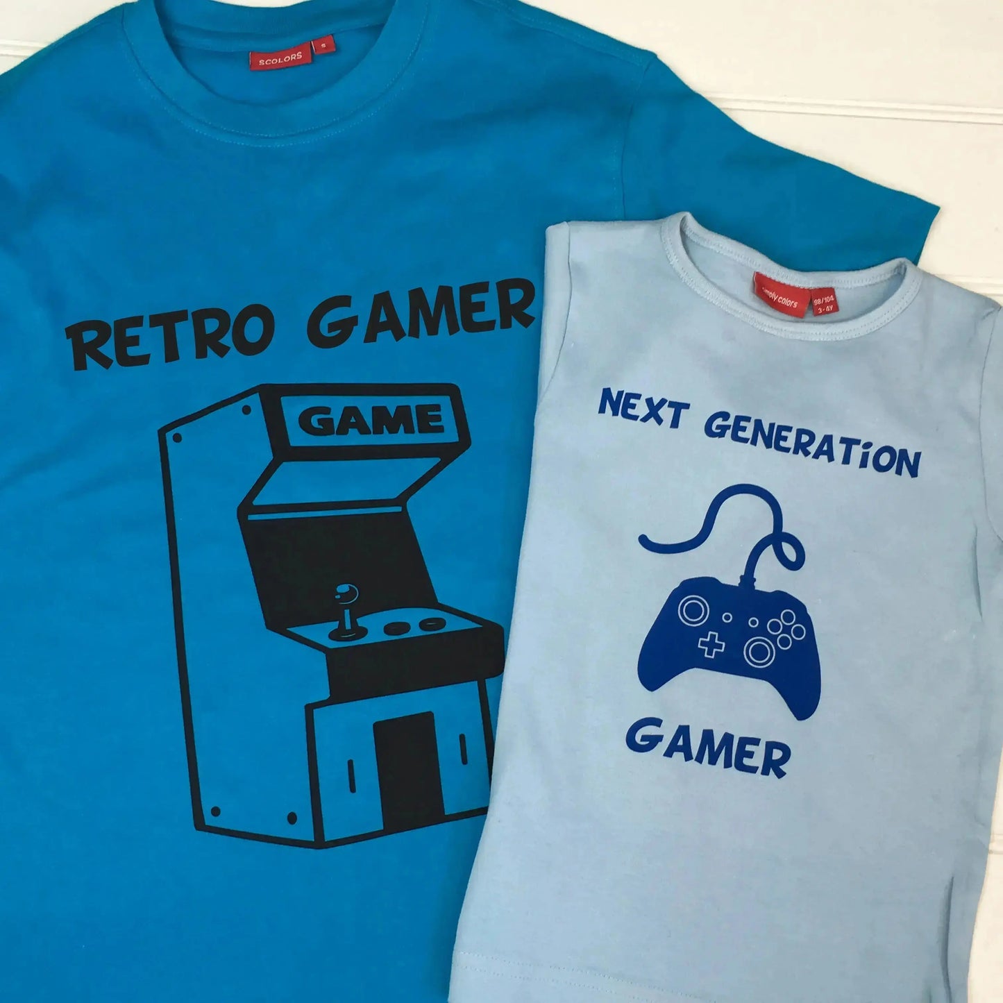 Adult t shirt with retro games console printed in black with Retro Gamer printed on top. Kids light blue t shirt with controller 1 printed in royal blue with two lines of text, top Next Generation, bottom line Gamer.