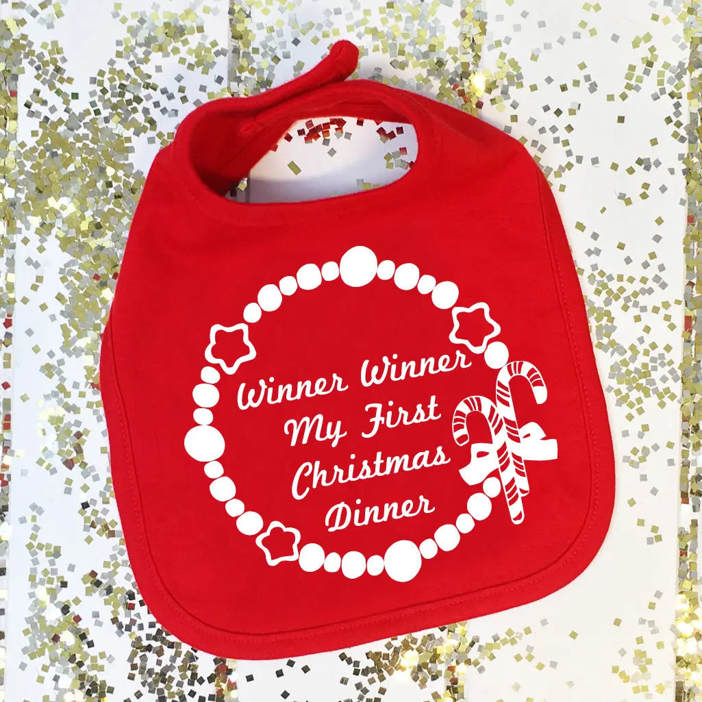Red bib with a circular Christmas design and text in the center that reads ' Winner Winner My First Christmas Dinner ' over four lines.