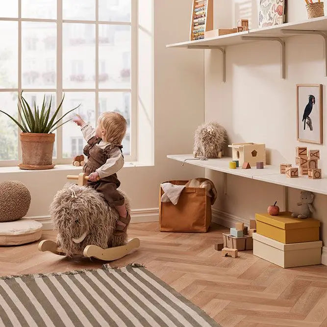 Child sitting on a rocking mammoth toy in a play room. She is looking out the large window and pointing. In the background is a pull along mammoth and other toys.