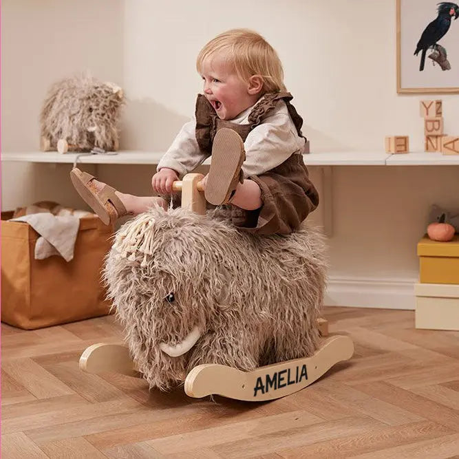 Little girl sitting on a personalised rocking mammoth toy, her name Amelia is printed on the side of the wooden rockers. She looks like she is having great fun, her legs are swinging up. The mammoth has scruffy fur, tassels on the top of his head for hair and white tusks along side his trunk. In the background is a pull along mammoth and some other toys.