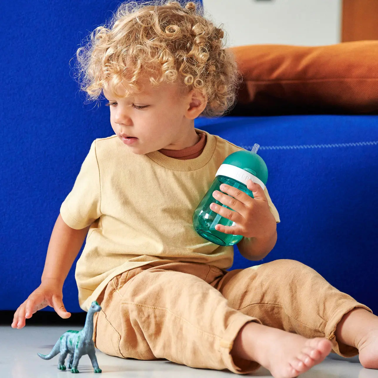 Toddler holding his straw water cup, the lid is open so the straw can be seen. He is sitting on the floor, playing with a dinosaur