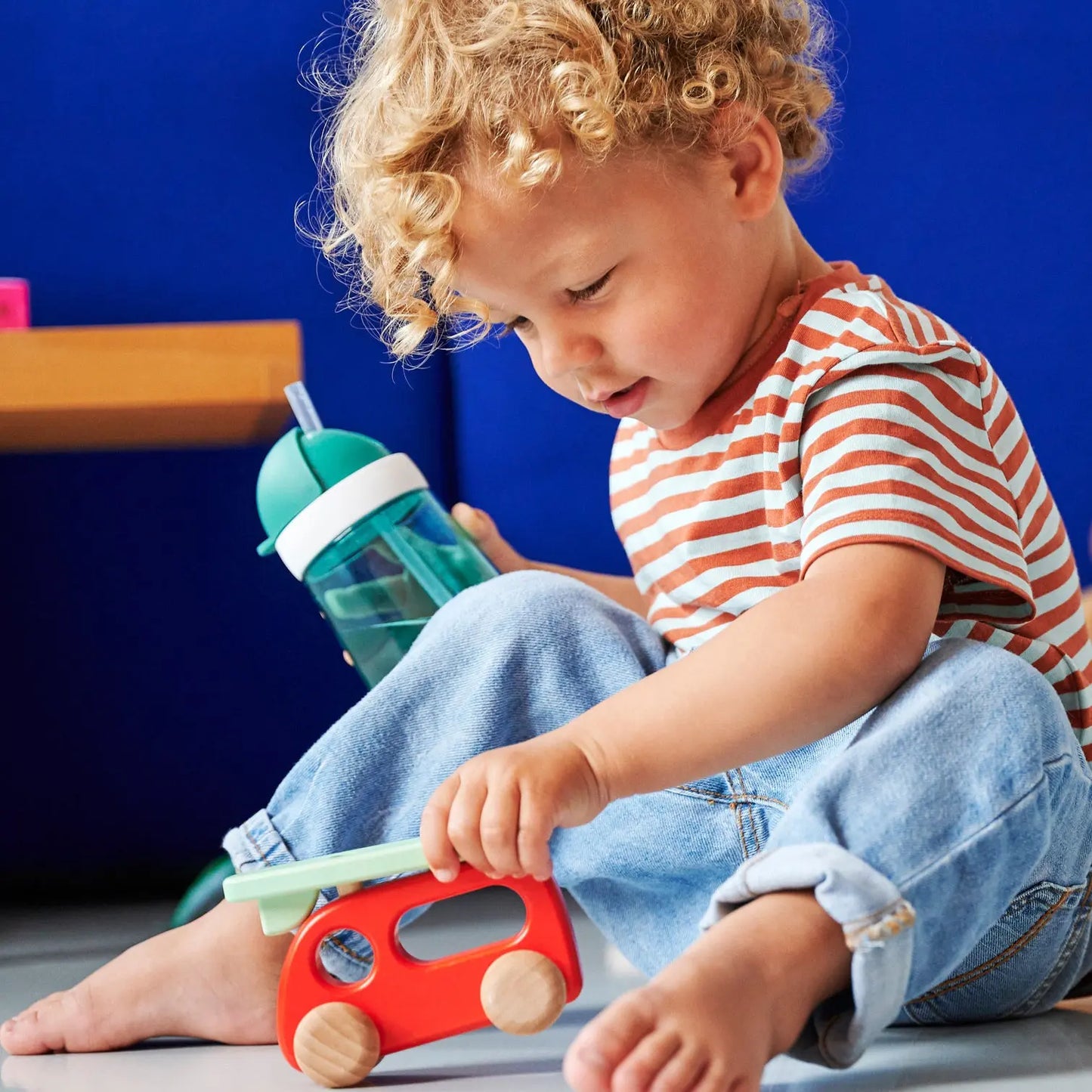 Toddler holding his turquoise straw water cup, with the lid open and the straw up ready to drink from. He is playing with a wooden car toy