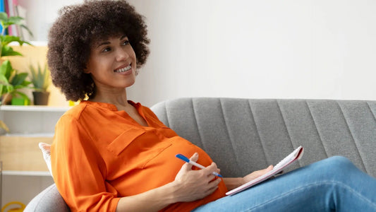 Pregnant lady on a sofa with a notepad and pen, thinking and smiling about baby names