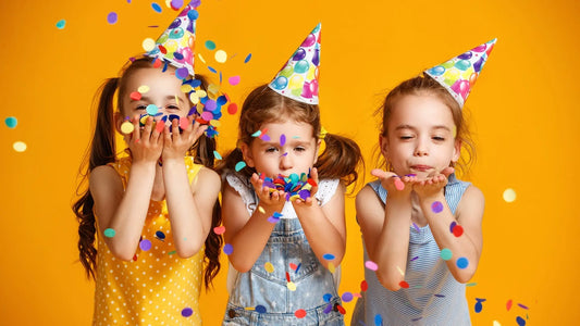 3 girls holding round colourful confetti and blowing it. They are wearing colourful party hats