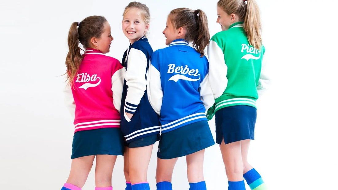 4 girls wearing varsity jackets printed with their names on the back. One wears pink, the others wear navy, blue and green.