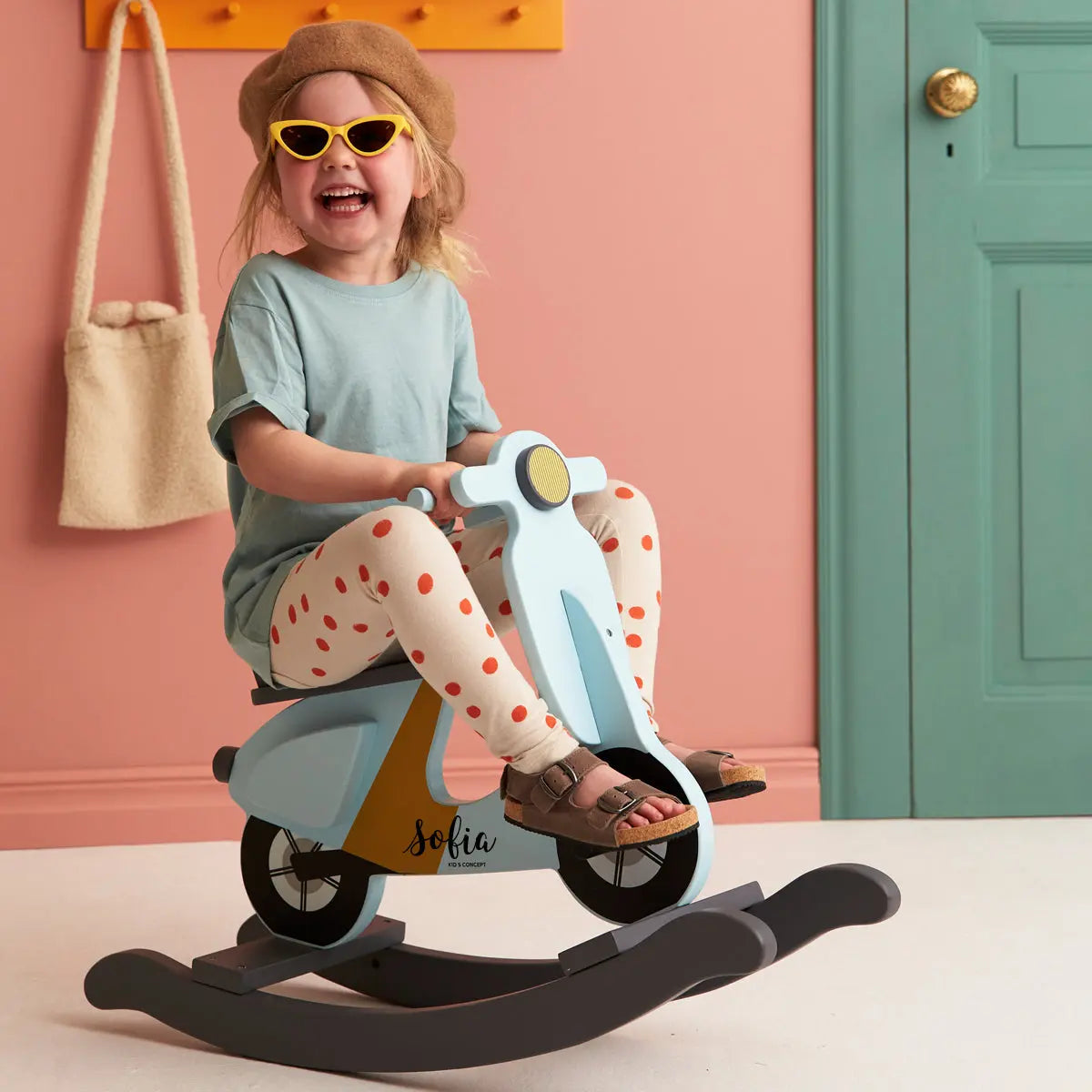A little girl playing on a blue rocking scooter with the name Sofia, She is smiling and wearing sun glasses