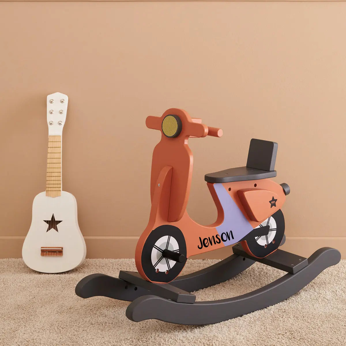 Personalised rocking scooter with the name Jenson in black, the scooter is rust and lilac. Next to the scooter is a kids white toy guitar.