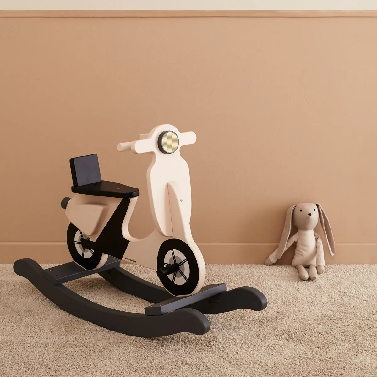 Light beige rocking scooter with a black seat and rockers. Next to the scooter is a bunny with very long ears.