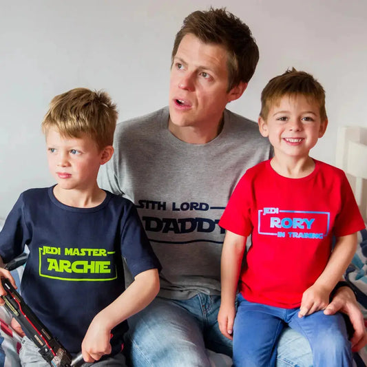A man and two children wearing star wars design t shirts. Dad wears Sith Lord Daddy, one son wears Jedi Master Archie and the other son wars Jedi in Training Rory.