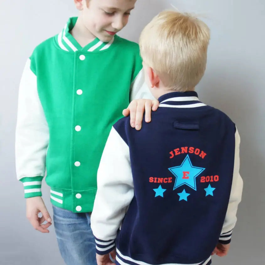 Two brothers wearing varsity style jackets. The youngest wears a navy jacket with white sleeves, printed on the back is a sporty star design with the name Jenson since 2010 and an E in the centre of the large blue star. There are 3 stars underneath the main star. The older boy wears a green varsity jacket with white sleeves, he is looking at his younger bother and has his hand resting on the brothers shoulder. The green jacket is buttoned up and shows the front of the jacket when closed.