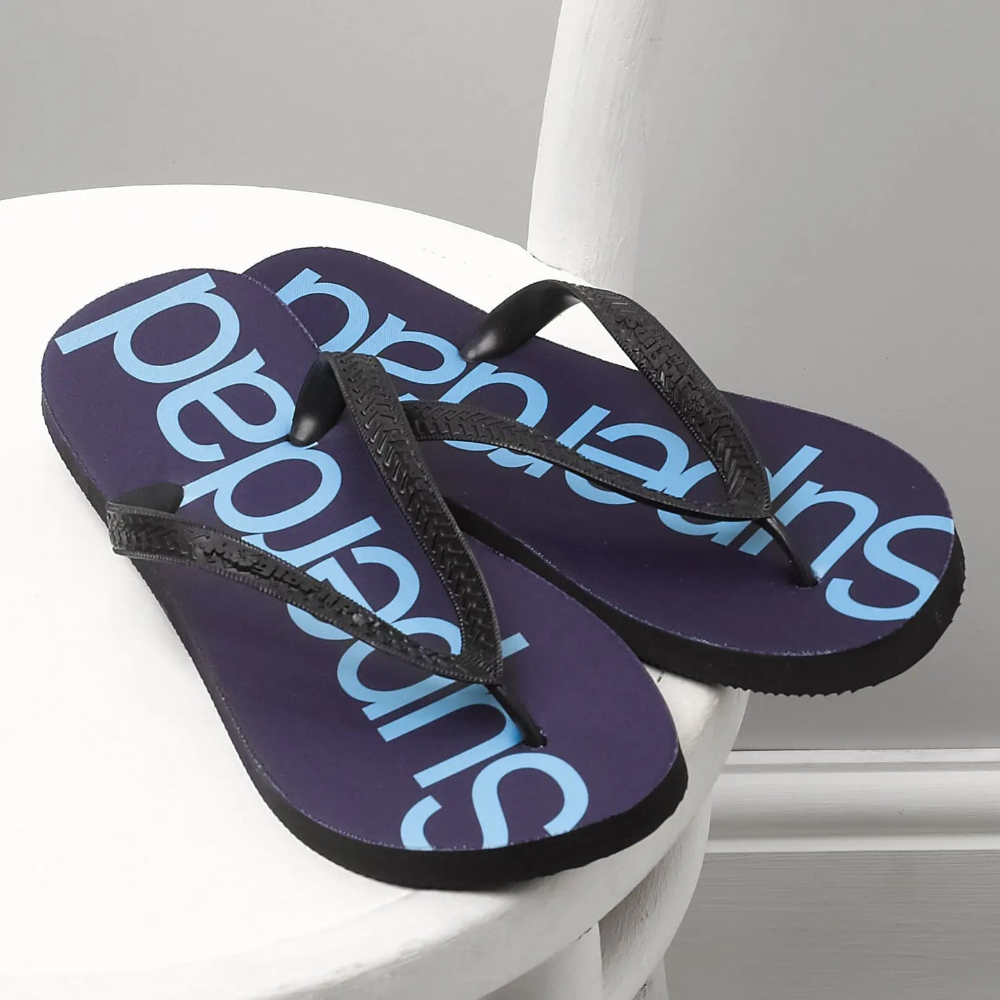 Pair of Superdad flip flops on a chair, the superdad is printed in light blue on navy background