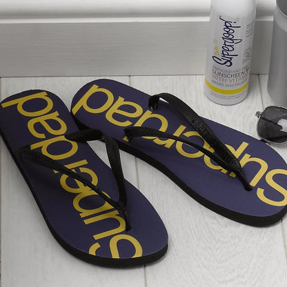 A pair of flip flops with Superdad printed down the centre in yellow, the base of the flip flop is navy. The is a bottle of suntan lotion and sun glasses next to the flip flops