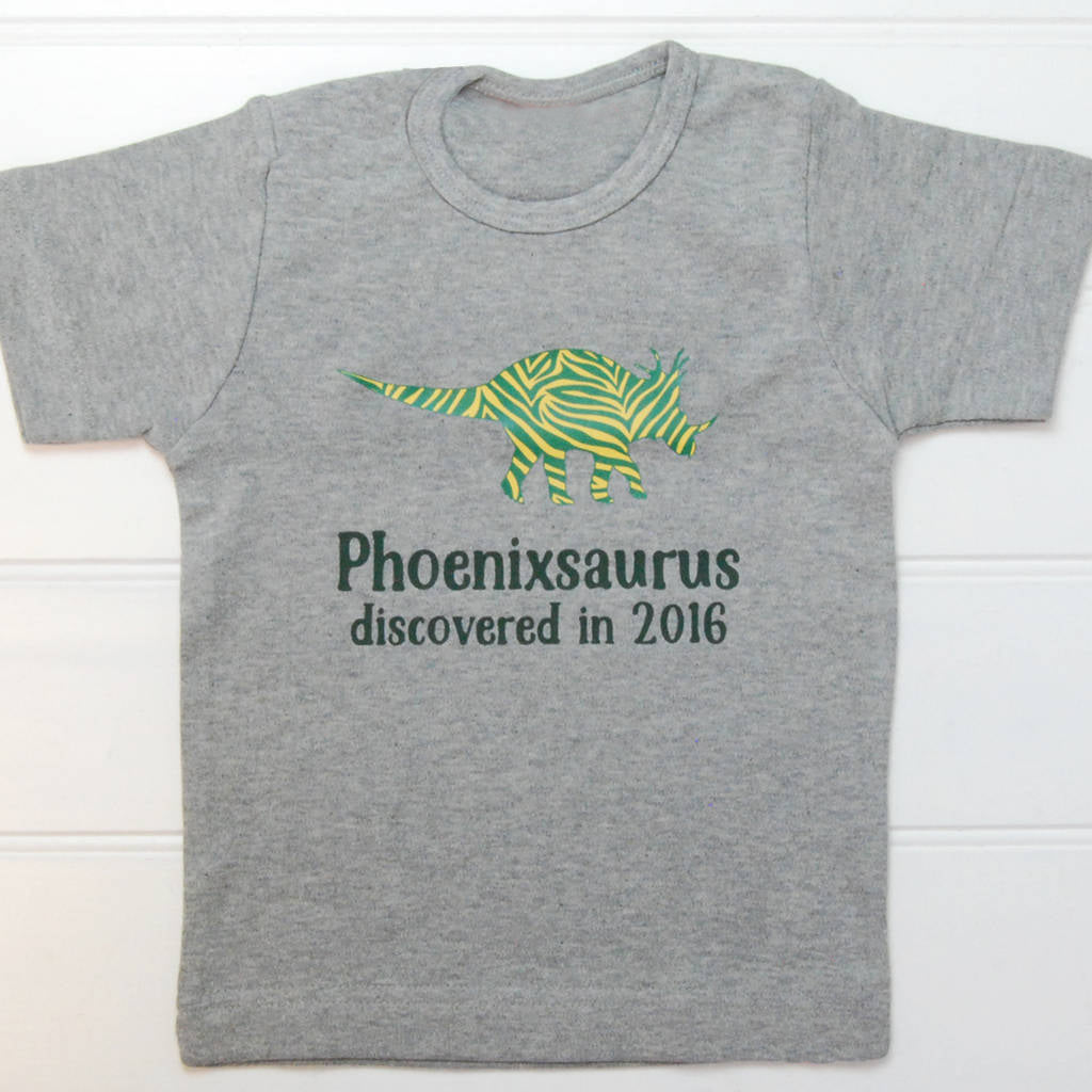 Grey t shirt with a green and yellow striped triceratops with the name Phoenixsaurus discovered in 2016 underneath printed in dark green on the front