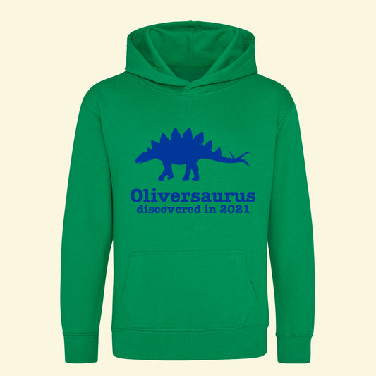 Green kids hoodie with stegosaurus printed on the front between the neck hole and top of the front pocket. Printed with two lines first line - Oliversaururs second line - discovered in 2021. The whole design is printed in royal blue.