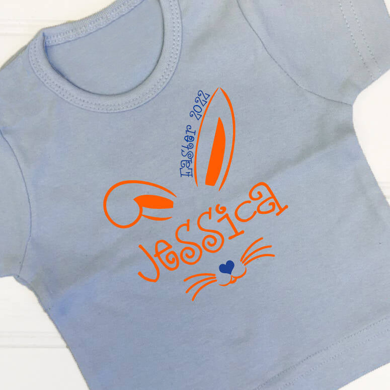 Children's light blue t shirt with an Easter bunny design. The name Jessica has bunny ears above and a cute nose with a heart and whiskers printed in neon orange. The text Easter 2022 is printed above one of the ears in royal blue, as is the heart. A very cute t shirt for Easter