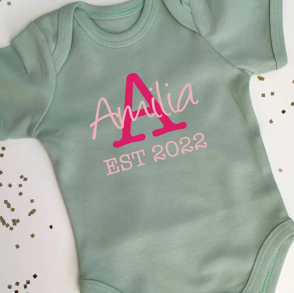 Light green babygrow with a capital A in fuchsia pink and the name Amelia in light pink over the centre of the A. Underneath is EST 2022 in light pink. This design is on the front of the babygrow