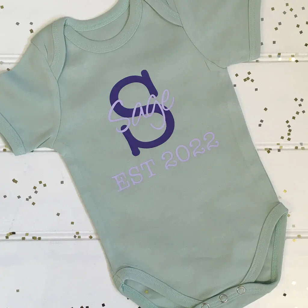 Light green babygrow with a capital S in purple with the name Sage printed in lilac over the centre and EST 2022 underneath on the front of the babygrow. The background is white with gold sprinkles.