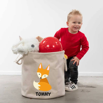 Little boy smiling next to a cotton storage bag that is filled with toys. The storage bag has the image of a fox and the name Tommy printed underneath.