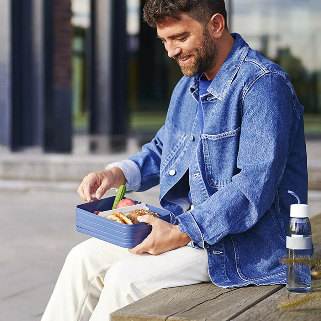 Man sitting with a large to go lunch box and ellipse water bottle. The lunch box has a divider in, separating his sandwiches and fruit