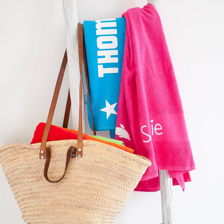 Pink and aqua blue beach towels printed with names and icons, hanging up with a beach bag