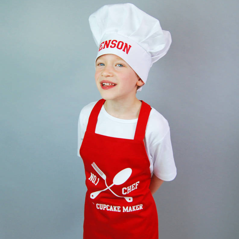 Boy wearing a red apron with a white t shirt. The apron has a knife and fork design with the text No. 1 Chef Chief Cupcake Maker printed on the centre front. The boy is also wearing a White chef hat with the name Jenson printed in red on the front band