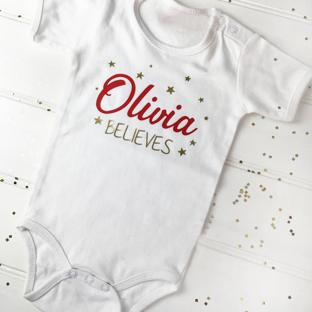 Cute white babygrow with a Christmas design. The text reads Olivia and then Believes under the name, with different size stars around the text. The name is printed in red and Believes and the stars in glitter gold