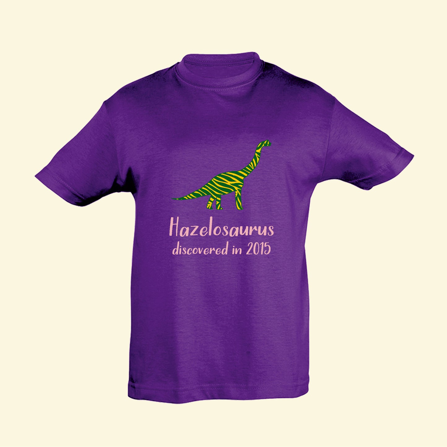 Purple t shirt with a brachiosaurus with a yellow and green stripe design. Underneath the name Hazelosaurus and discovered in 2015 printed in light pink on the front.