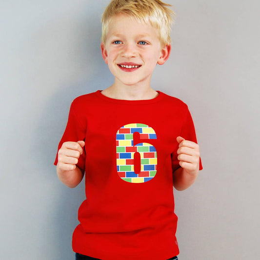 Boy wearing a red t shirt with the number 6 printed on the front. The number has a lego brick style design with vibrant prime colours. Ideal birthday t shirt