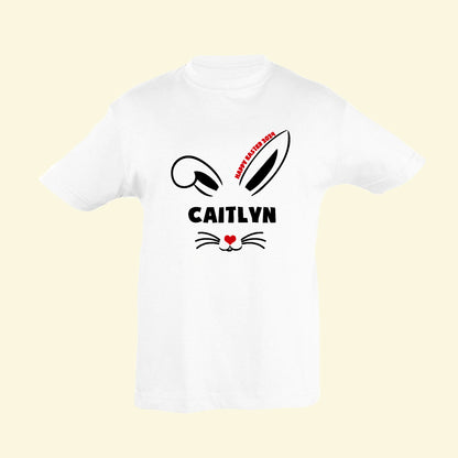 White t shirt with a bunny design printed on the front. The cute bunny ears have Happy Easter 2024 along side one of them, then the name Caitlyn and below the name a nose with whiskers and a heart in the centre. Perfect to wear on Easter Egg hunts