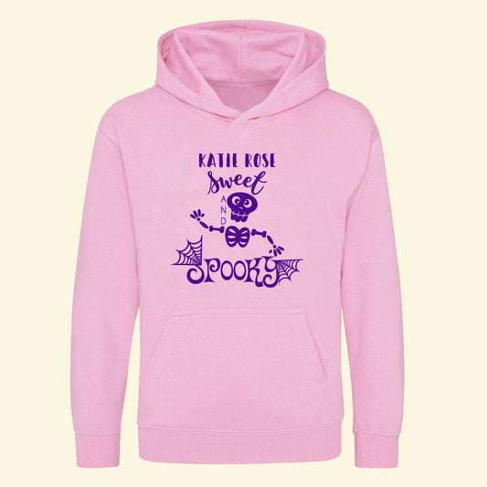 Light pink hoodie with a halloween design printed on the front. The design has a skeleton, cobwebs and the text Sweet and Spooky, along side the child's name printed in purple