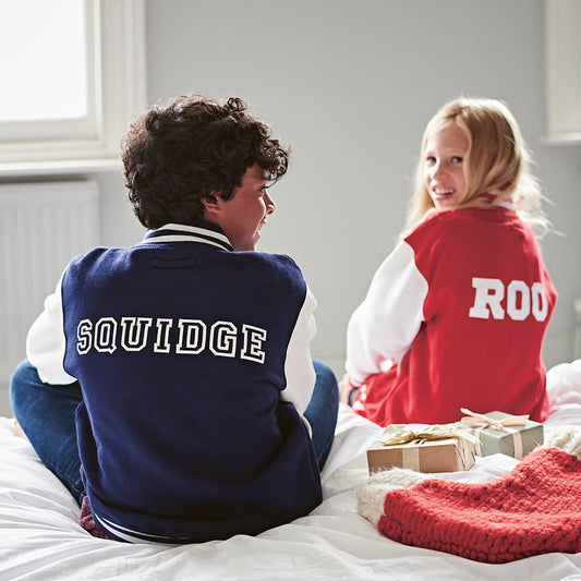 Two children wearing personalised varsity jackets, a boy wearing a navy jacket with the name Squidge on the back and a girl wearing a red jacket with Roo printed on the back. The children are sitting on a bed with presents and a Christmas stocking