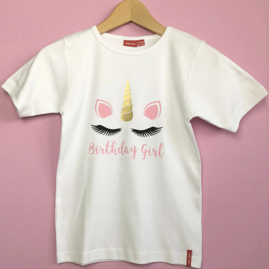 Kids white t shirt with Birthday Girl printed below black eyelashes, golden horn in the middle of two light pink ears matching the text.