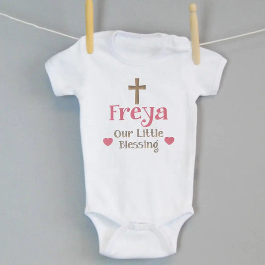 White babygrow with a traditional cross and the text Freya, Our little Blessing with two hearts either side printed in gold and light pink on the front of the babygrow