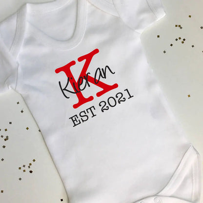 White babygrow with a large capital K in red with the name Kieran in black over the middle of the K, underneath in black is EST 2021. Surrounding the babygrow is a white background with gold sprinkles